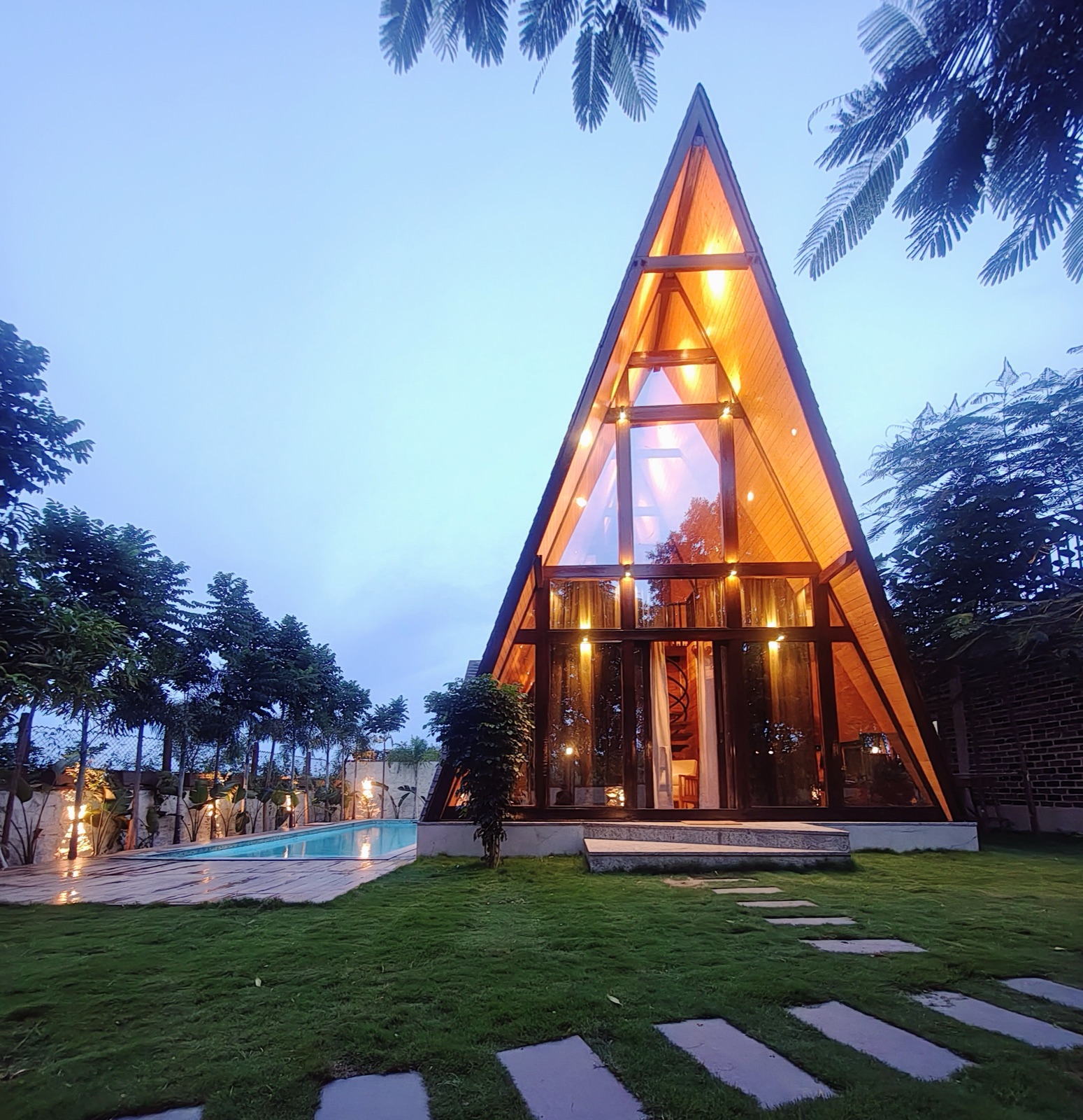 Glowing timber A-frame cabin beside a private swimming pool at dusk, framed by lush green trees