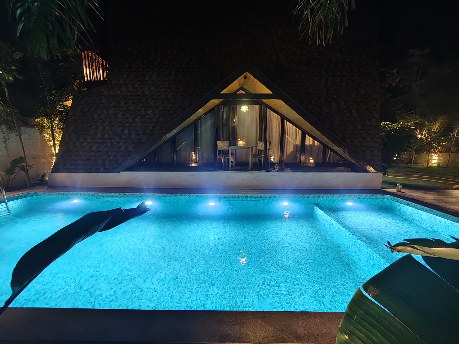 The A-frame cabin glowing with warm light beside the blue-lit pool at night